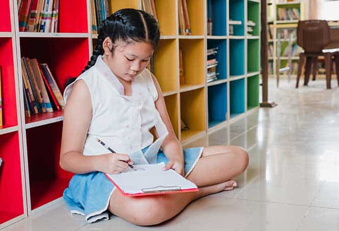 Girl sitting on the floor reading a book in a library setting.