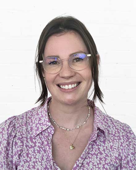 Woman with glasses, necklace, and purple patterned blouse smiling at the camera.